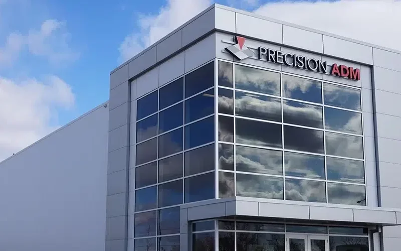 A modern industrial building with large security glass windows, reflecting clouds in the sky. The sign Precision ADM is mounted on the corner, showcasing Winnipegs architectural prowess. The sky is partly cloudy.