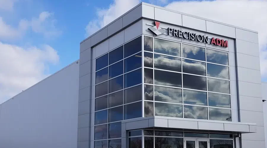 A modern industrial building with large security glass windows, reflecting clouds in the sky. The sign Precision ADM is mounted on the corner, showcasing Winnipegs architectural prowess. The sky is partly cloudy.