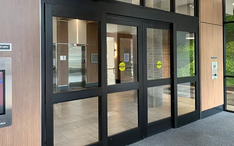 The entrance boasts security glass sliding doors, with a visible directory guiding visitors. Inside, the tiled floors and wooden walls complement green decor on the right. An elevator in the background is partially open, hinting at Winnipegs seamless blend of style and function.