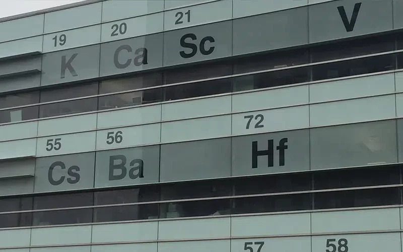 A building facade in Winnipeg, designed to resemble the periodic table, showcases elements with atomic numbers 19 (K), 20 (Ca), 21 (Sc), 55 (Cs), 56 (Ba), and 72 (Hf). Large security glass windows sit between the labeled panels, blending style with protection.