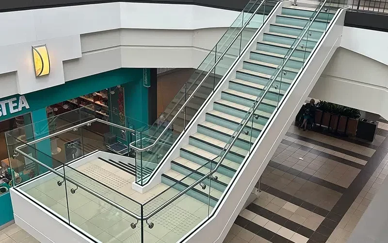 A modern indoor mall scene features a wide staircase with sleek security glass railings. On the left, a teal-colored storefront displays a partially visible logo and cozy seating area. The floor is tiled in a beige and brown grid pattern, reminiscent of Winnipegs contemporary design aesthetics.