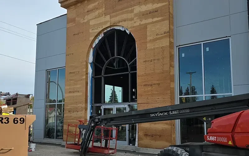 A building under construction with a large arched entrance being framed features Security Glass Winnipeg windows. A red Skyjack lift is parked in front, and a wooden panel covers part of the facade, while the reflective windows capture the surroundings beautifully.