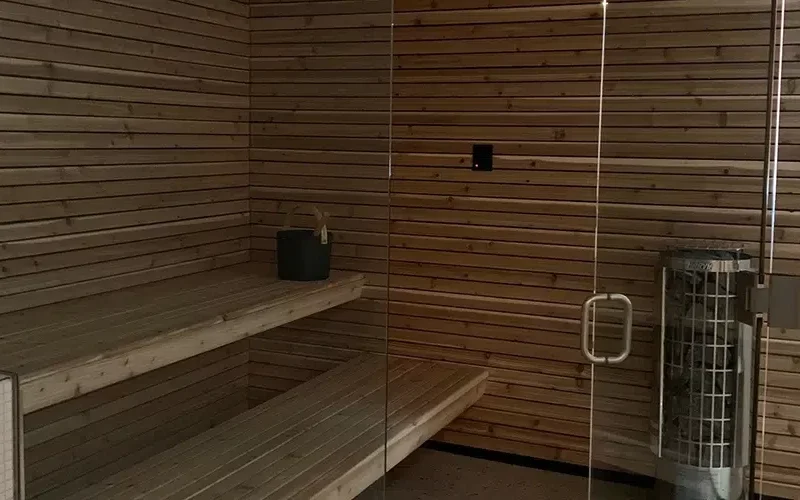 A wooden sauna room with two-tiered seating benches on the left and a heater on the right. The walls and ceiling are crafted from horizontal wooden slats, while a sturdy security glass door from Winnipeg stands in the foreground, providing both safety and style.