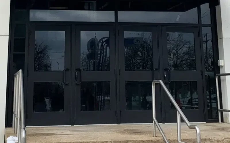 Glass double doors at the entrance of a building, featuring security glass from Winnipeg, reflect trees and the street outside. A stone staircase leads up to the doors with metal railings on both sides. A red fire alarm pull station is visible on the right.