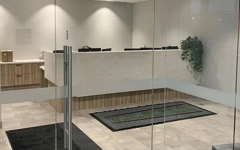 A modern office reception area features a wooden and marble front desk. A potted plant adorns the right side, and glass doors, equipped with Security Glass Winnipeg, display a TEAMS logo on the floor mat. The tiled floor is in a light gray color.