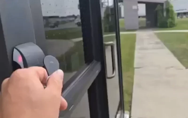 A hand holding a key card is about to tap it on a security glass card reader mounted beside the glass door. Outside, a sidewalk leads to another building across the grassy area in Winnipeg, with a parked truck visible in the distance.